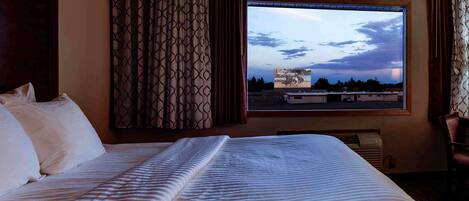A hotel room with a large bed, a window with a view of buildings, and a wall-mounted air conditioner.