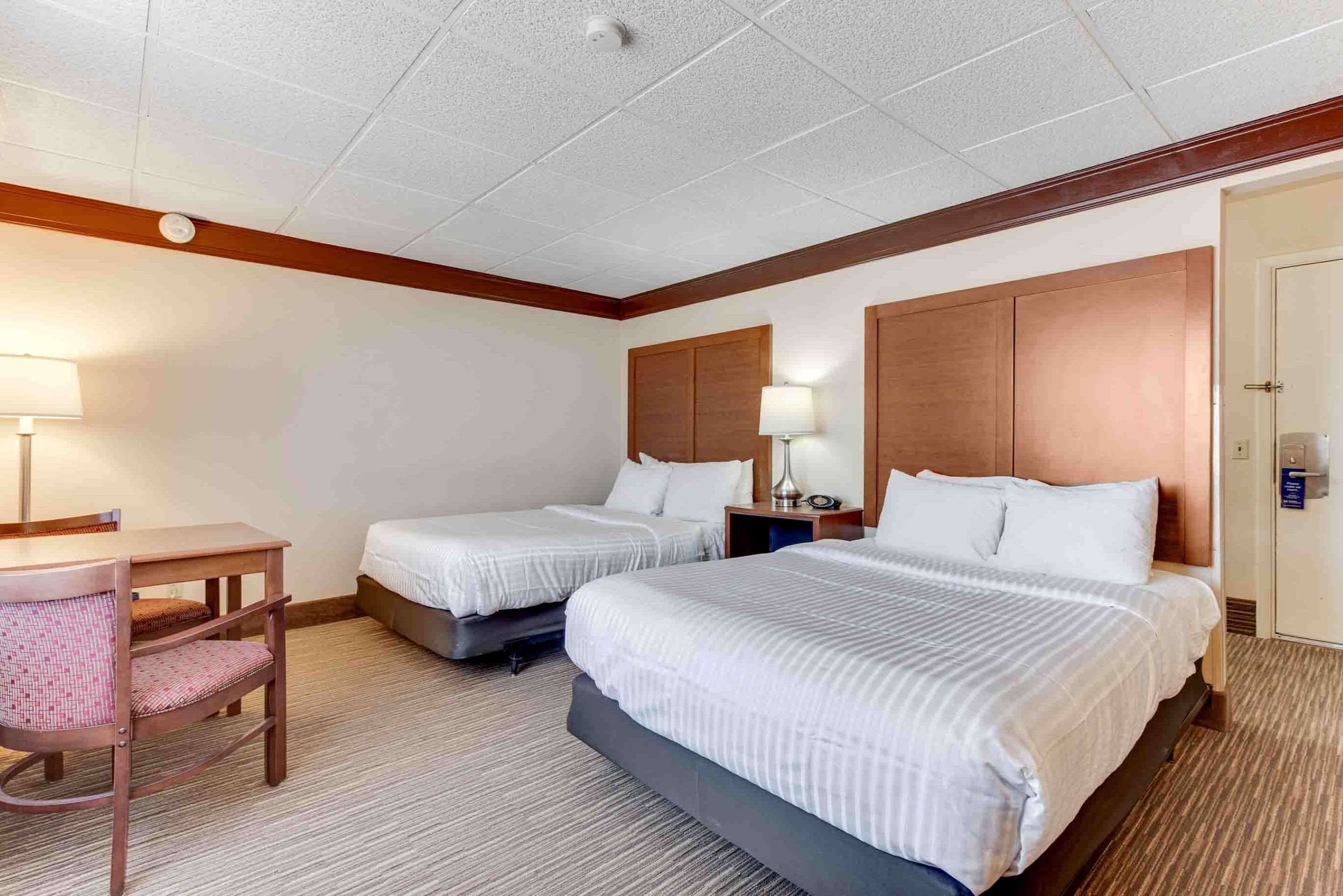Ein Hotelzimmer mit zwei Betten, einem Schreibtisch, einem Stuhl und einem Nachttisch mit Lampe.