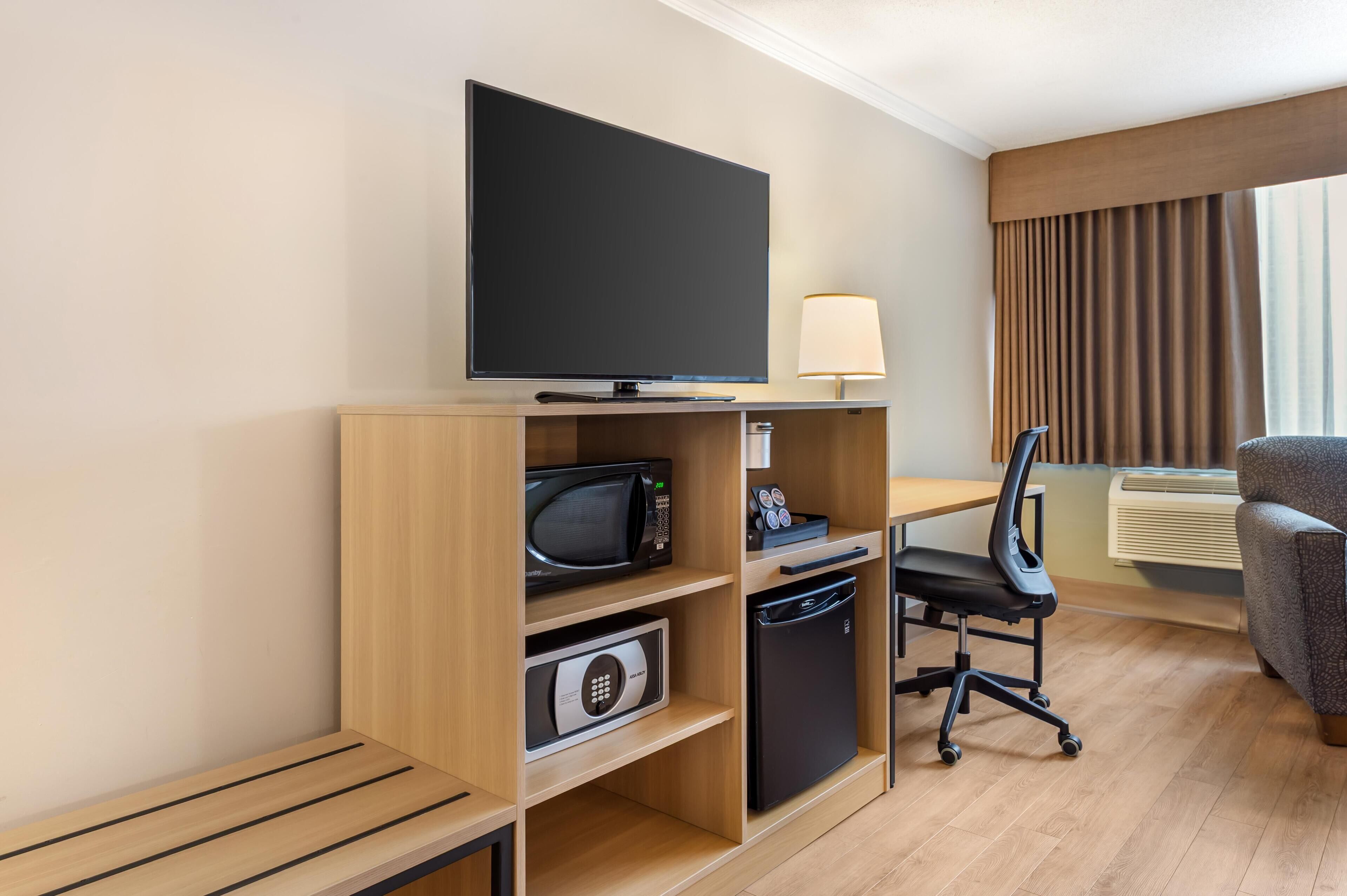 A hotel room with a flat-screen TV, a microwave, a mini-fridge, a desk with a chair, a lamp, and a large window with curtains.