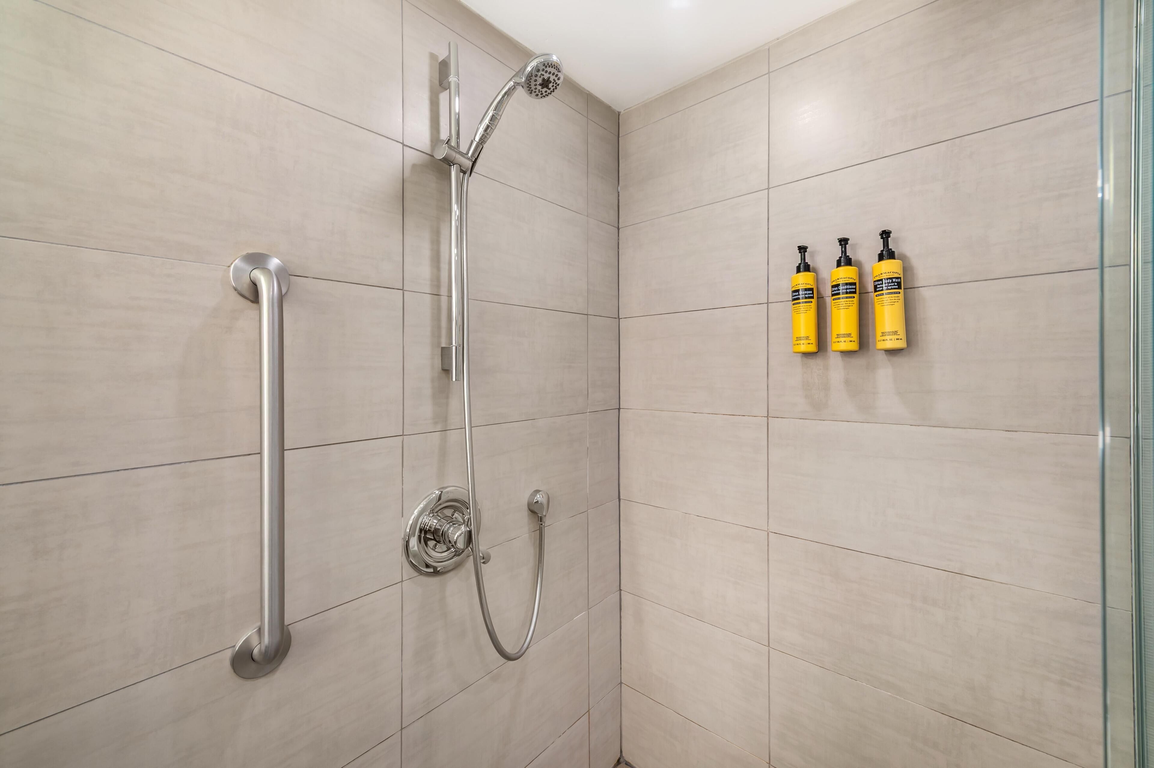 Zona de ducha con cabezal de ducha manual, cabezal de ducha fijo y manguera de ducha manual. Hay tres botellas amarillas de champú y acondicionador en la pared.