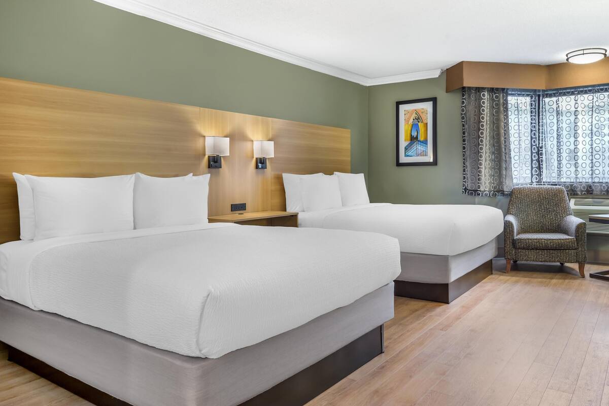 A hotel room with two beds, a wooden headboard, wall-mounted lamps, a chair, and a window with patterned curtains.
