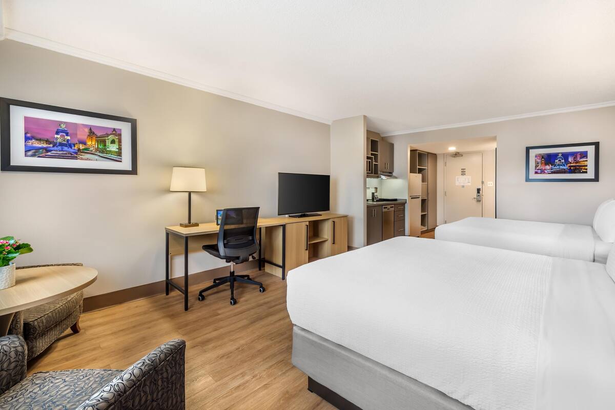 A hotel room with a bed, a desk with a computer and TV, a chair, a small table with a plant, and a picture on the wall.