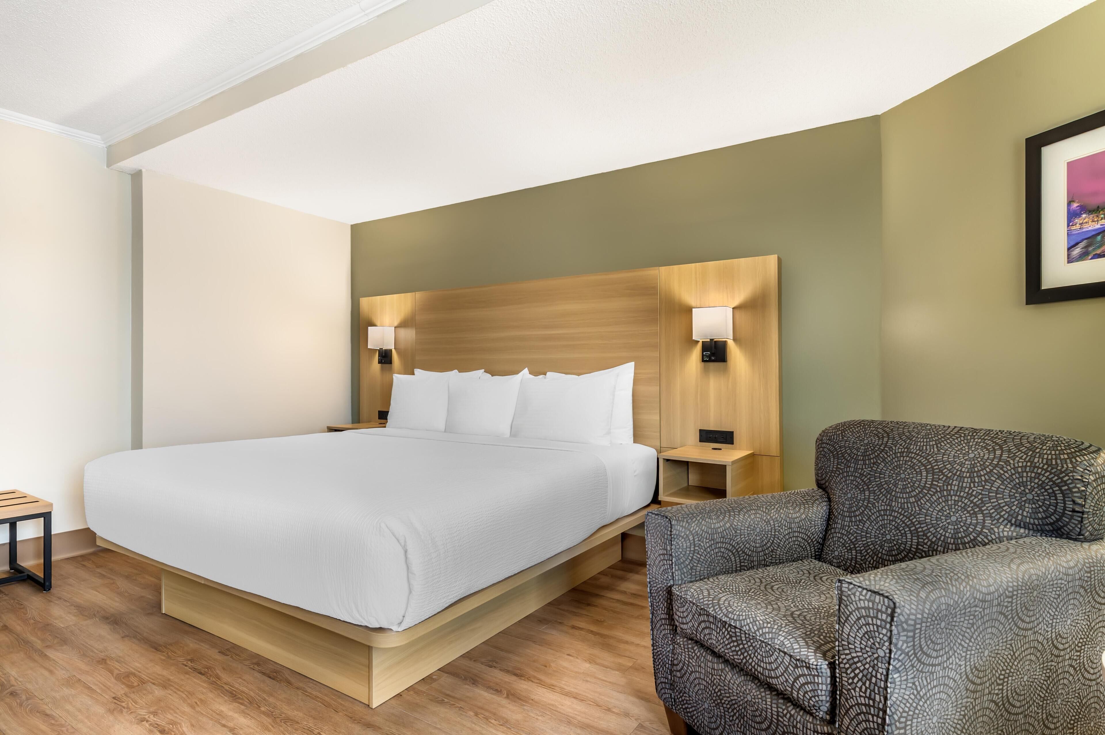 Une chambre d’hôtel moderne avec un grand lit, deux tables de chevet avec des lampes, un fauteuil à motifs et un tableau encadré au mur.