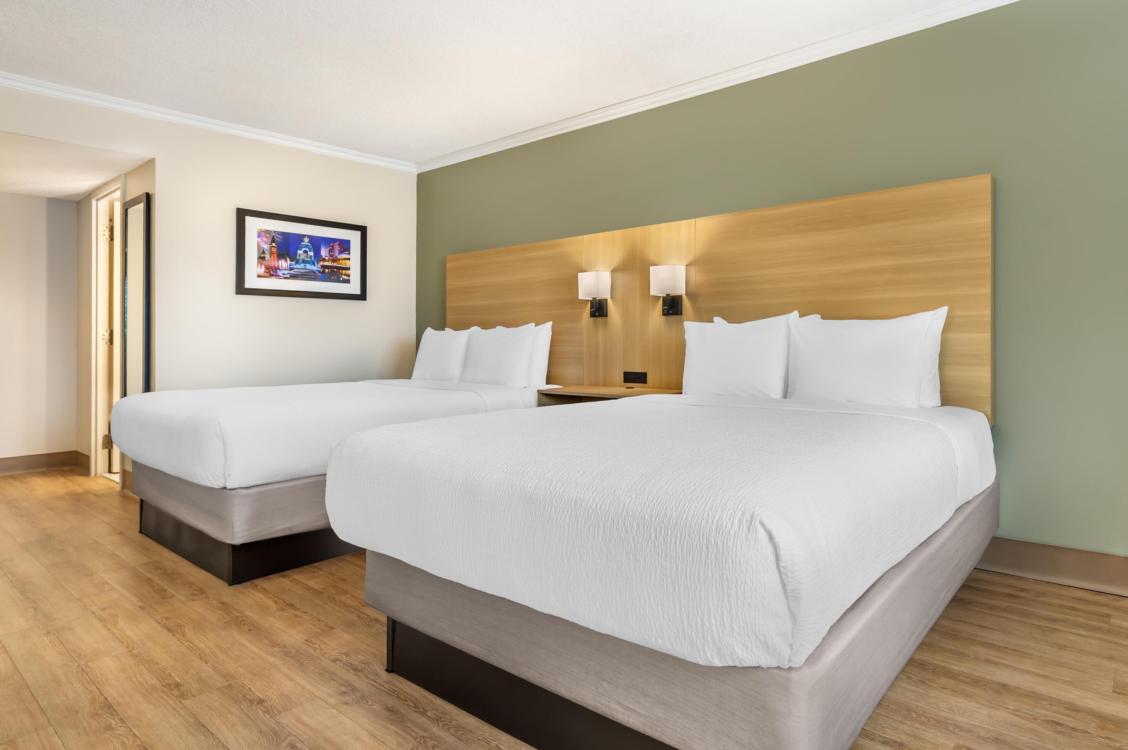 Une chambre d’hôtel avec deux lits, une tête de lit en bois, des lampes fixées au mur et un tableau encadré accroché au mur.