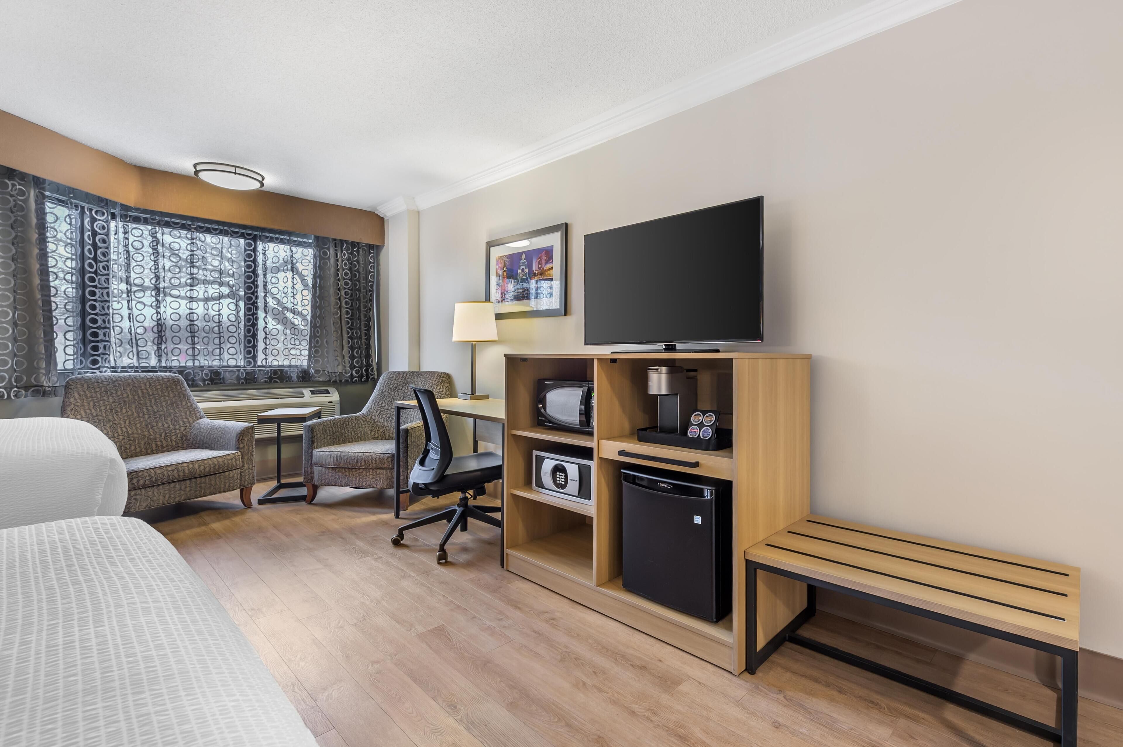 A hotel room with a bed, a desk with a computer, a chair, a TV, a microwave, a refrigerator, and a window with curtains.