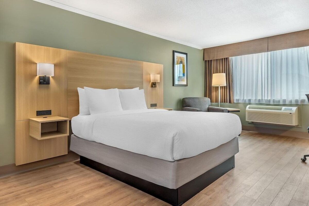 A hotel room with a large bed, bedside tables, a chair, a desk, a lamp, and a window with curtains.