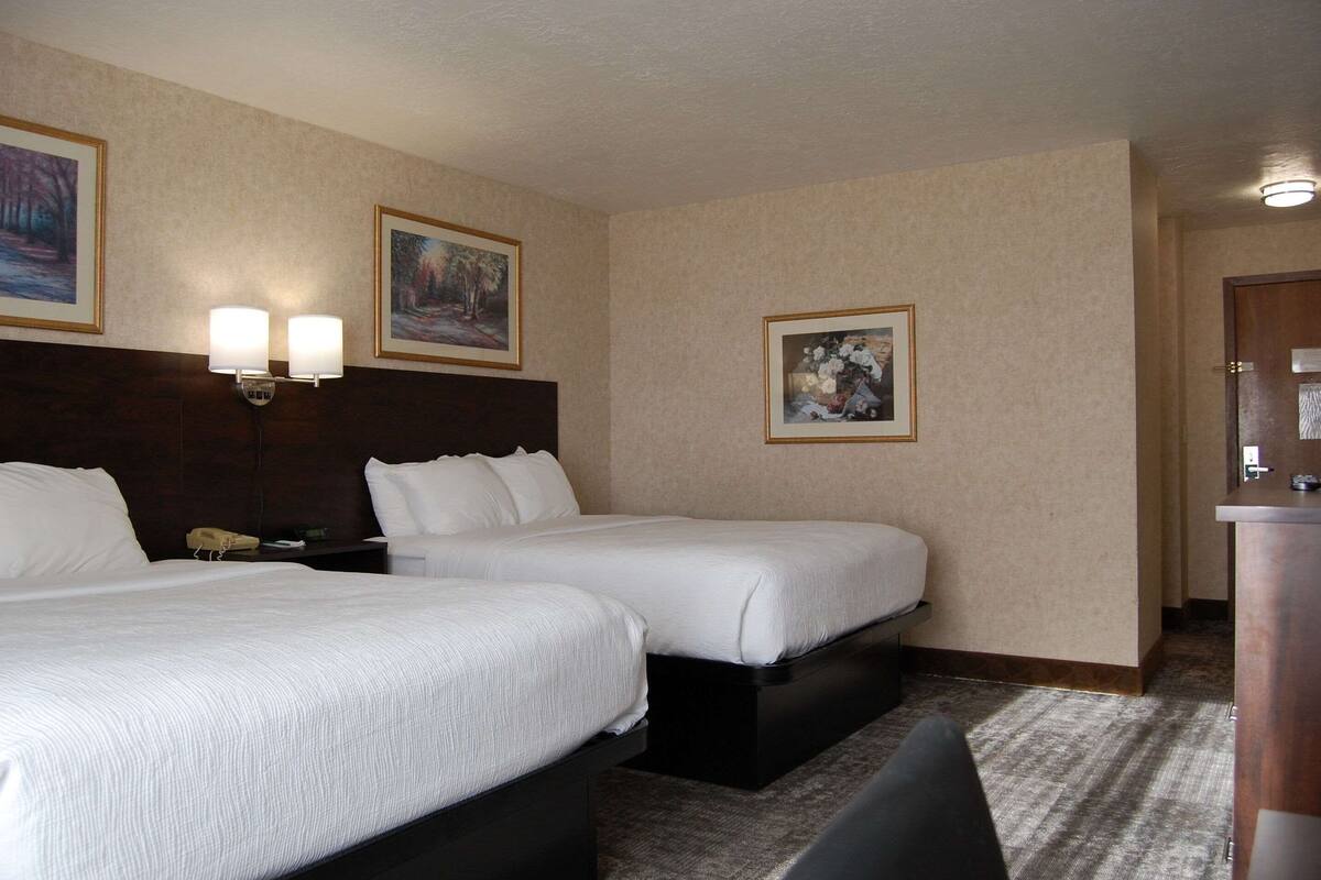 A hotel room with two beds, a desk, and a chair. There are framed pictures on the walls and a lamp on the desk.