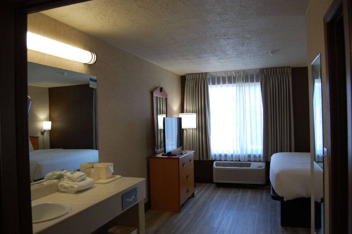 A hotel room with a bed, a desk with a computer, a mirror, a dresser, a bathroom with a sink and toilet, and a window with curtains.