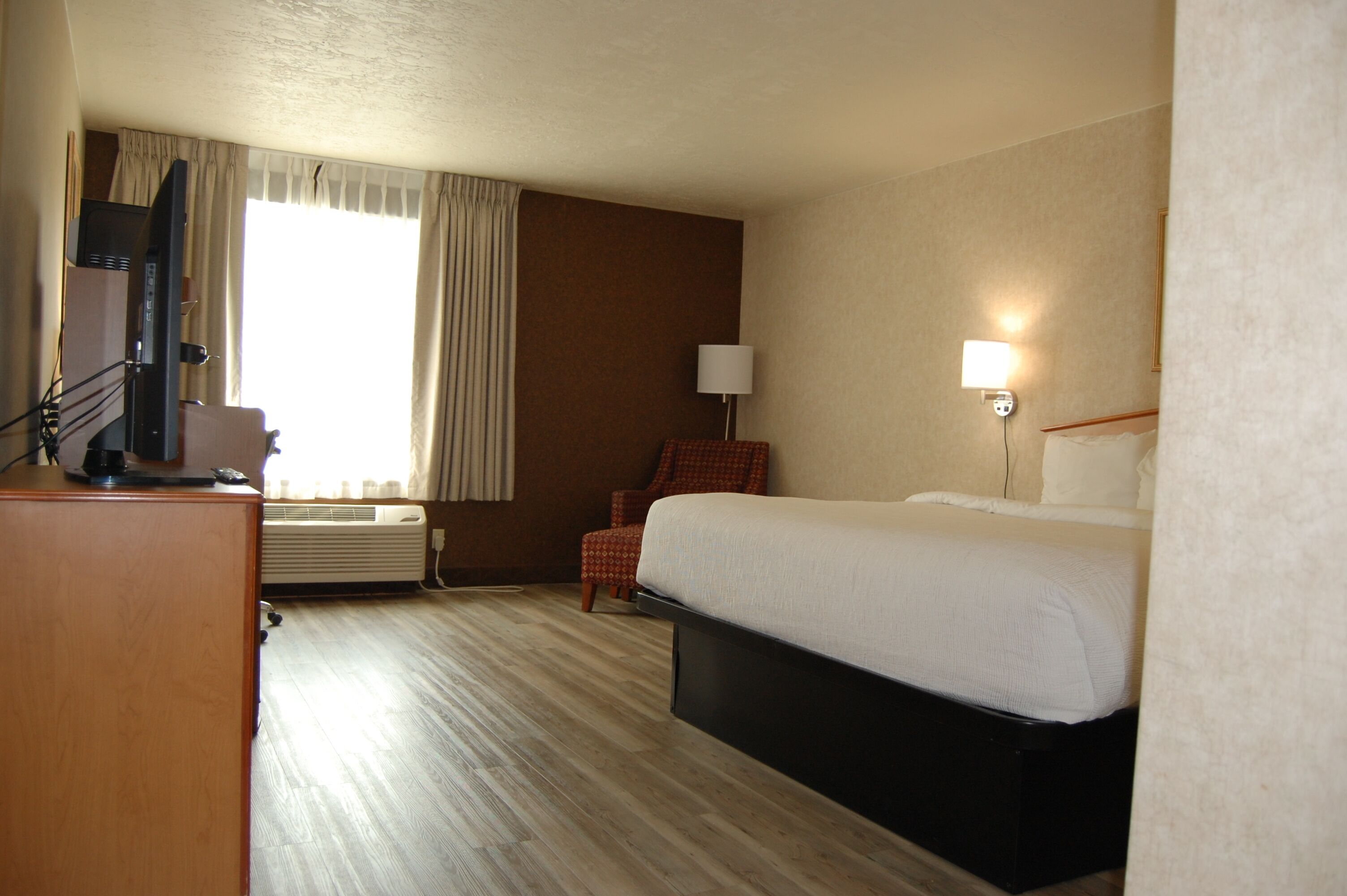 Una habitación de hotel con cama, escritorio con computadora, silla, lámpara y ventana con cortinas.