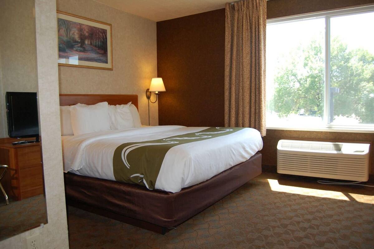 A hotel room with a bed, a TV, a lamp, a window with curtains, and an air conditioning unit.