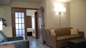 Hotellihuoneessa on ruskea nahkasohva, harmaa nojatuoli, puinen sohvapöytä ja yöpöytä. Suuresta ikkunasta avautuu näkymä ulkona olevaan rakennukseen.