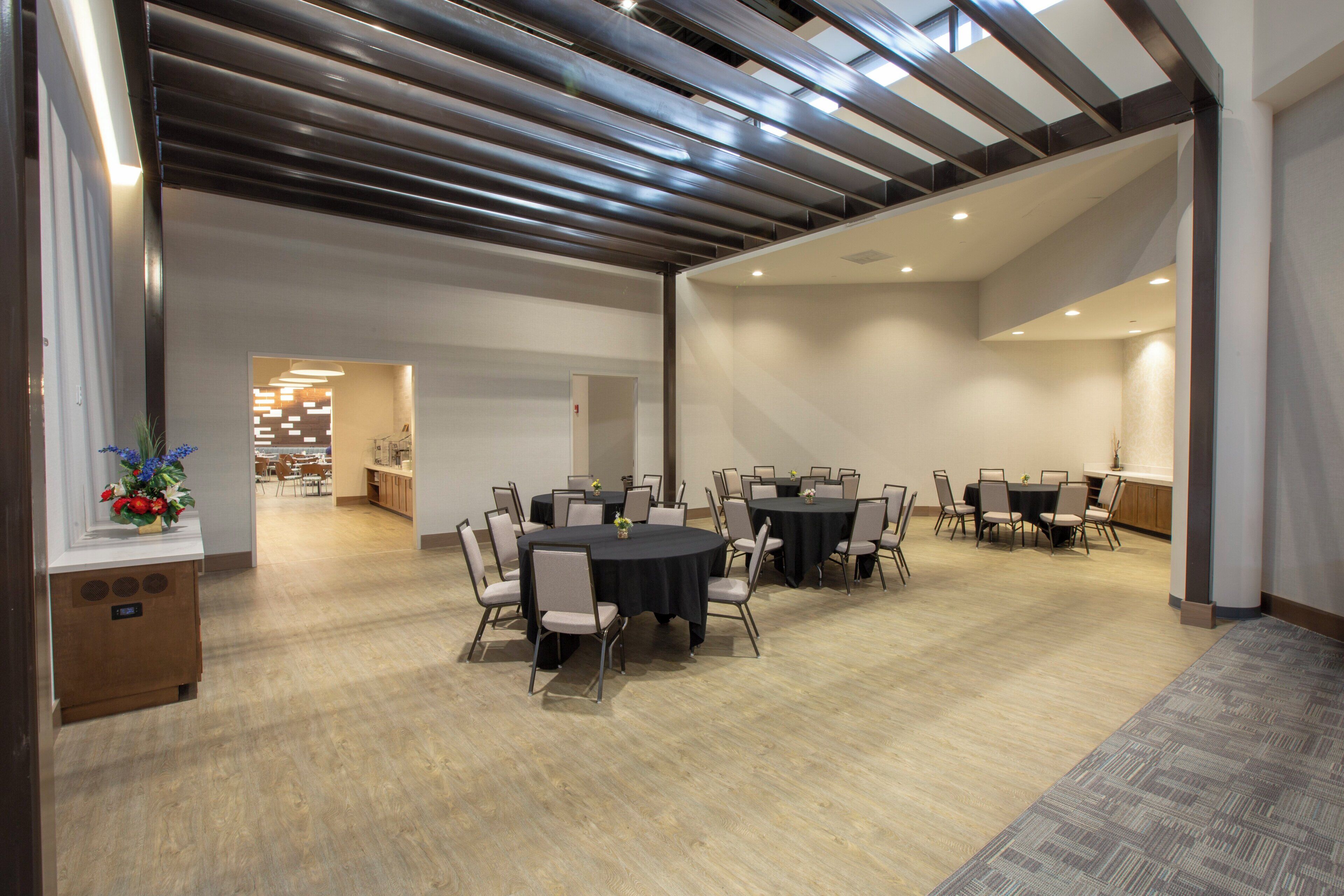 A spacious conference room with multiple tables and chairs, a modern ceiling design, and a visible kitchen area in the background.