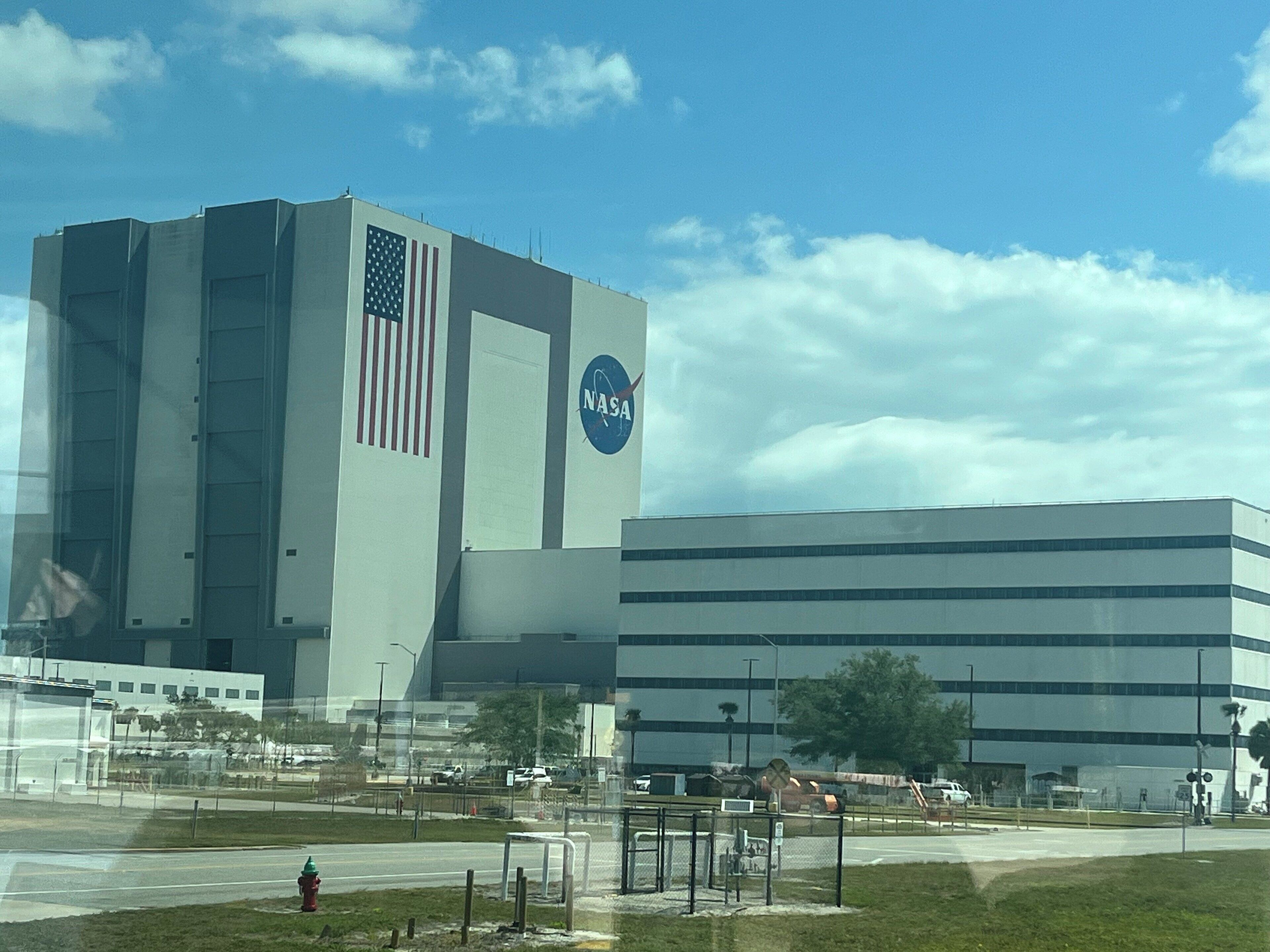 Un edificio grande con el logotipo de la NASA y una bandera estadounidense, rodeado de un estacionamiento y una zona verde.