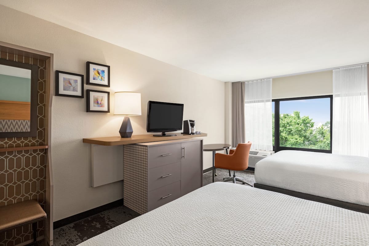 A hotel room with two beds, a desk with a television, a chair, and a window with a view of trees.