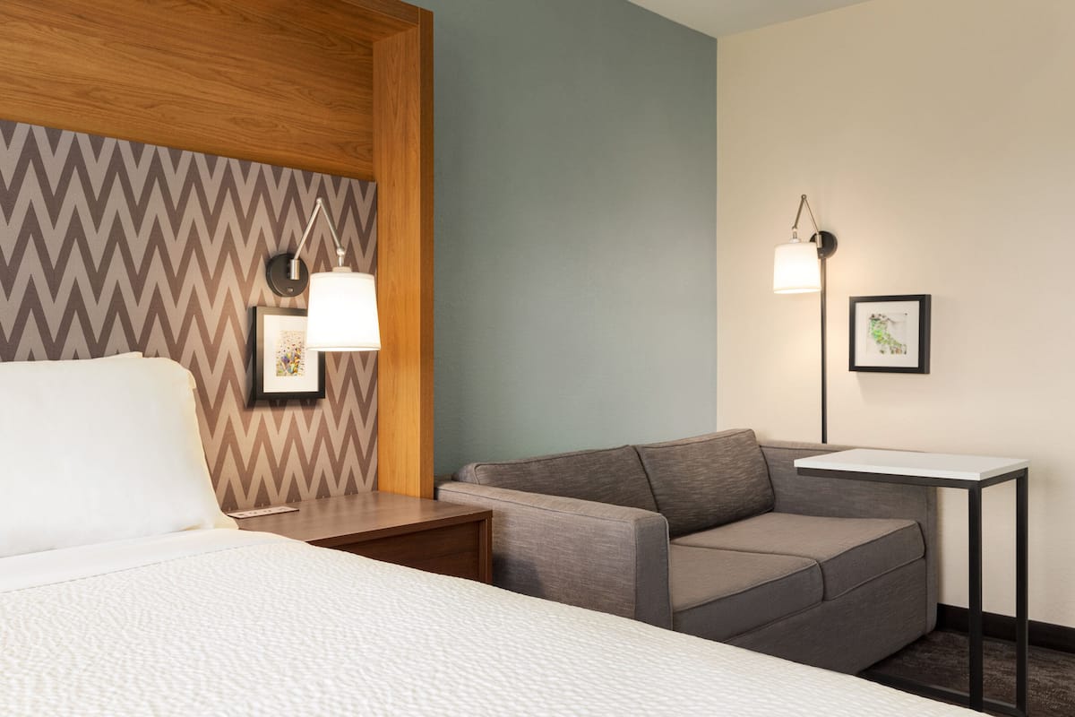 A hotel room with a bed, a gray sofa, a small table, and two wall-mounted lamps.