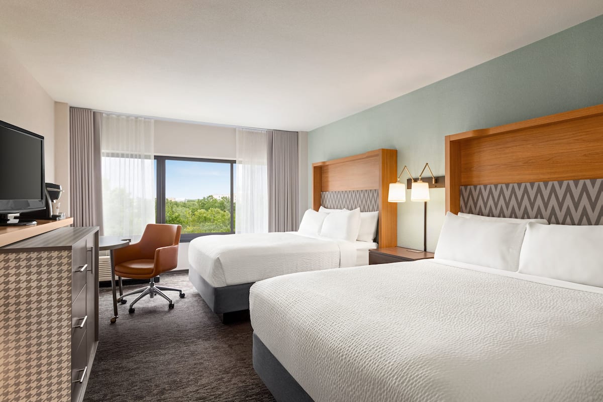 A hotel room with two beds, a desk with a TV, a chair, and a large window offering a view of greenery.