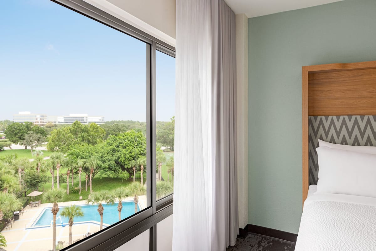 A hotel room with a large window overlooking a swimming pool and greenery, a bed with a wooden headboard, and a view of a building in the distance.