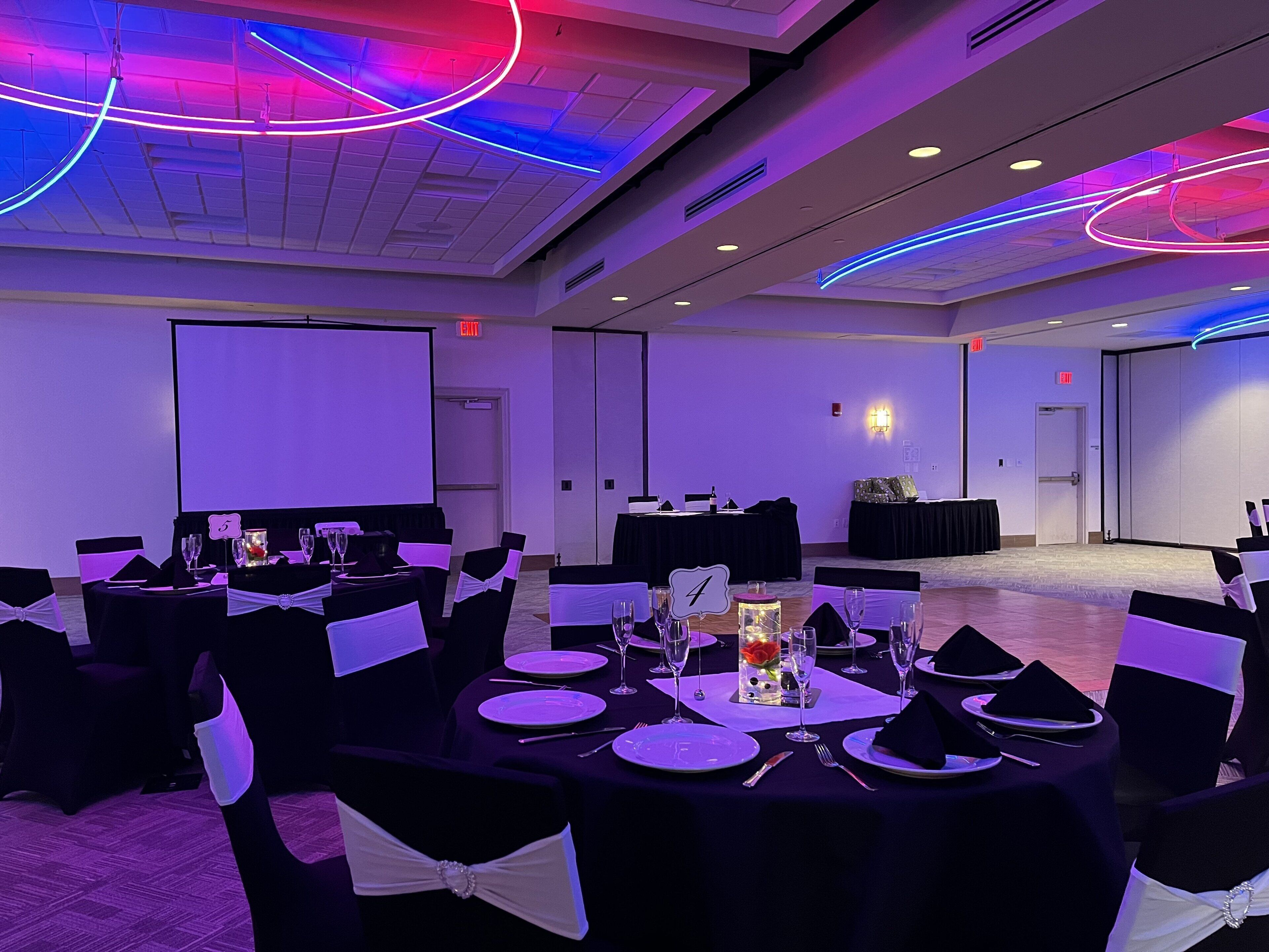 Un salón de conferencias con mesas dispuestas para un evento formal, equipado con proyectores y una pantalla grande.
