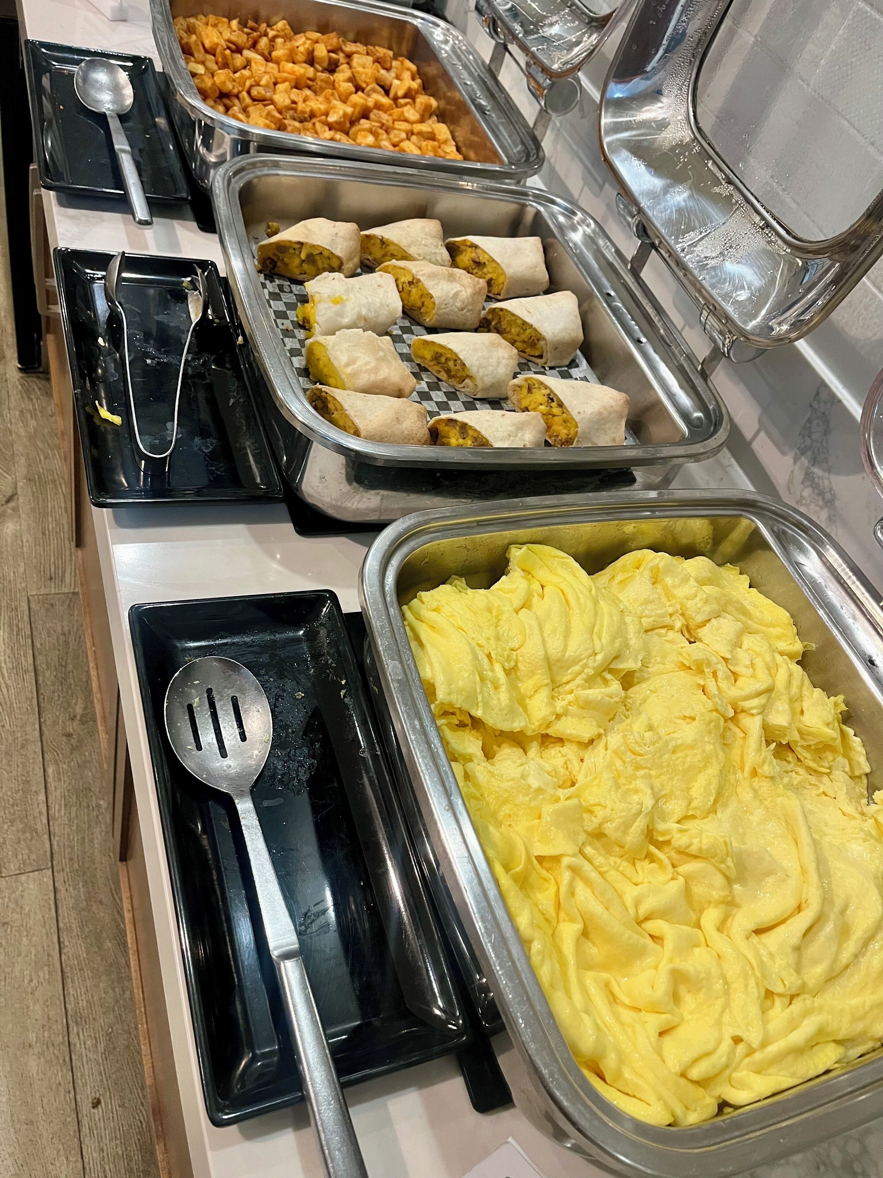 A buffet with various food items including burritos, a large container of scrambled eggs, and a tray of cubed food.