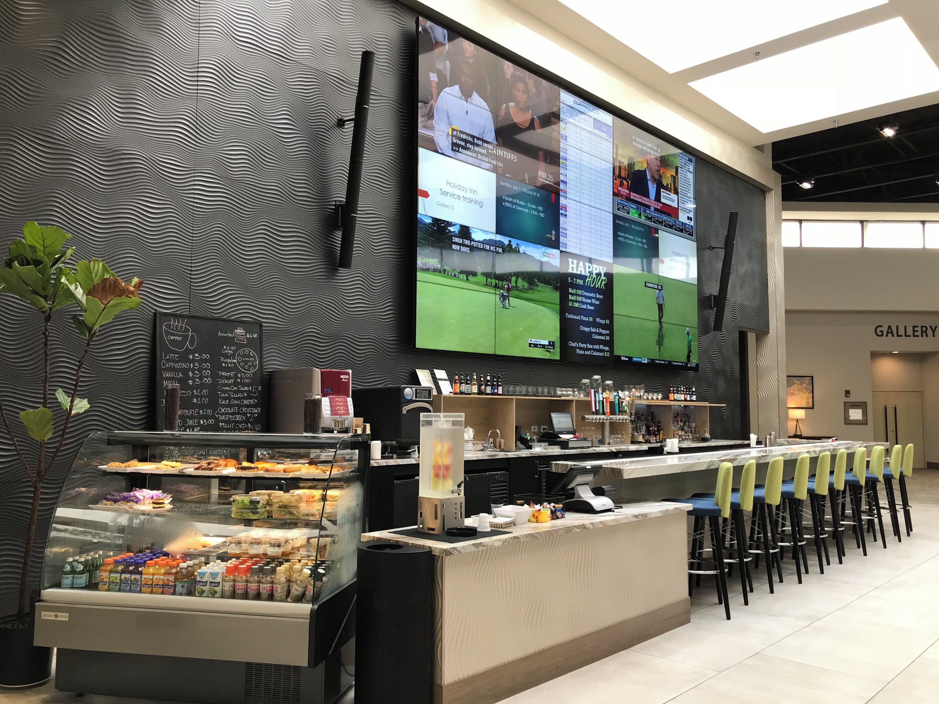 Un área de bar moderna con una gran pantalla mostrando deportes, una estación de café y un espacio de descanso con sillones.