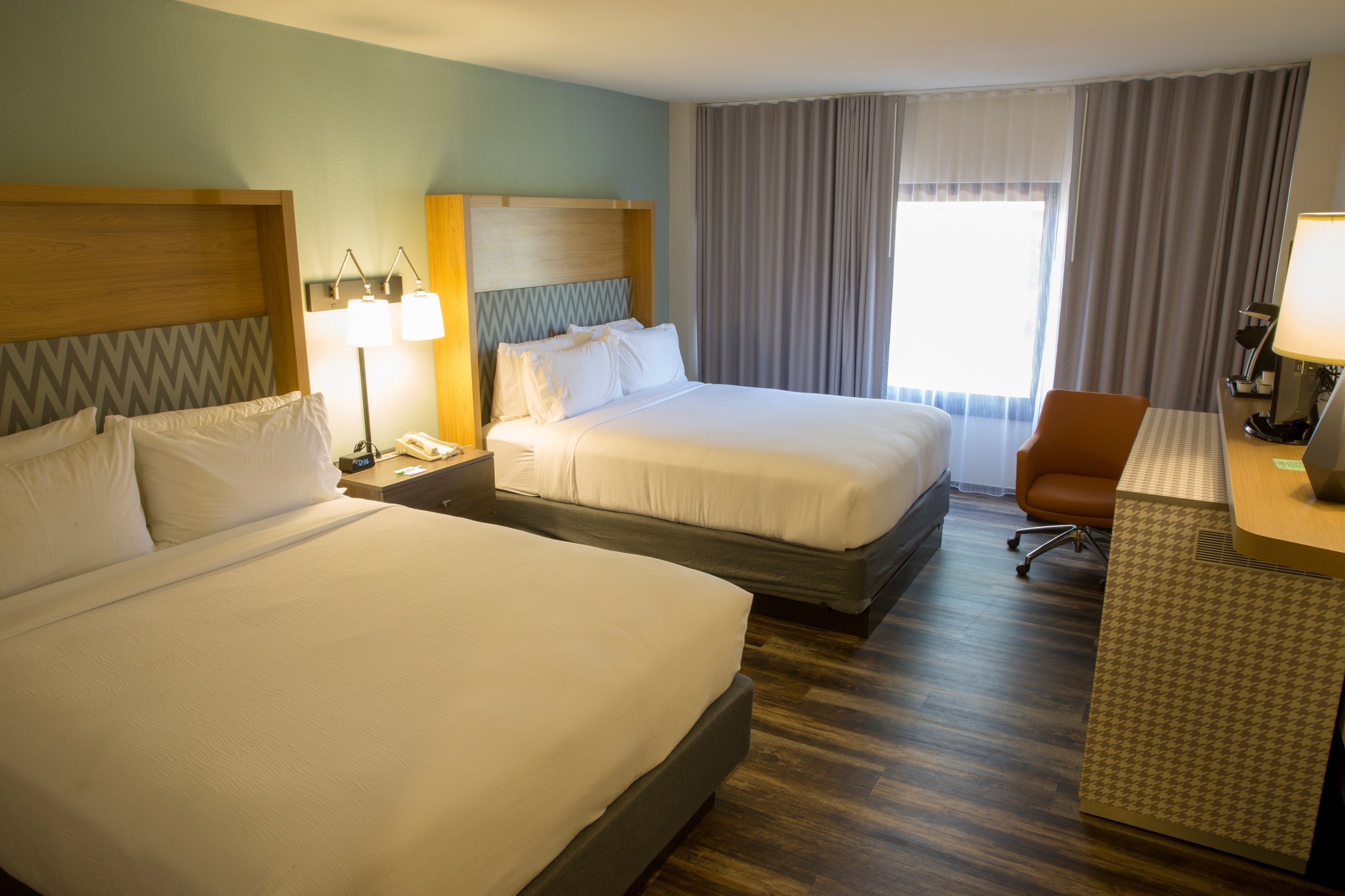 Habitación de hotel con dos camas grandes, un escritorio con silla, una lámpara y un ventanal con cortinas.