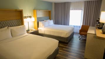 Habitación de hotel con dos camas grandes, un escritorio con silla, una lámpara y un ventanal con cortinas.