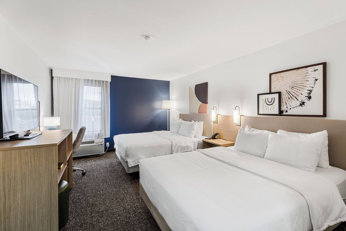 A hotel room with two beds, a desk, a chair, a desk lamp, a bedside table with a lamp, and artwork on the wall.