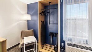 Une chambre d’hôtel avec un mur d’accent bleu, une armoire en bois, une table blanche, une chaise, un portant à vêtements et une fenêtre avec des rideaux légers.