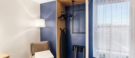 Habitación de hotel con pared azul decorativa, armario de madera, mesa blanca, una silla, perchero con ropa y ventana con cortinas traslúcidas.