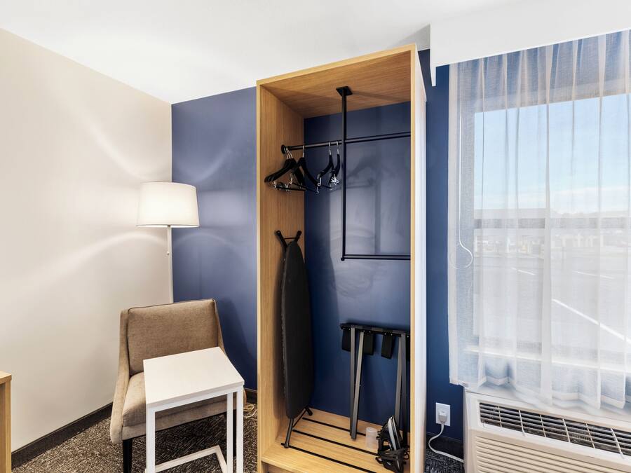 A hotel room with a blue accent wall, a wooden wardrobe, a white table, a chair, a hanging rack with clothes, and a window with sheer curtains.