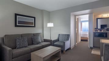 A hotel room with a grey sofa, a patterned armchair, a wooden coffee table, a floor lamp, a TV, and a microwave.