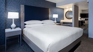 Moderni hotellihuone, jossa on suuri sänky, kaksi yöpöytää valaisimineen, pyöreä peili ja työpöytä tuolilla.