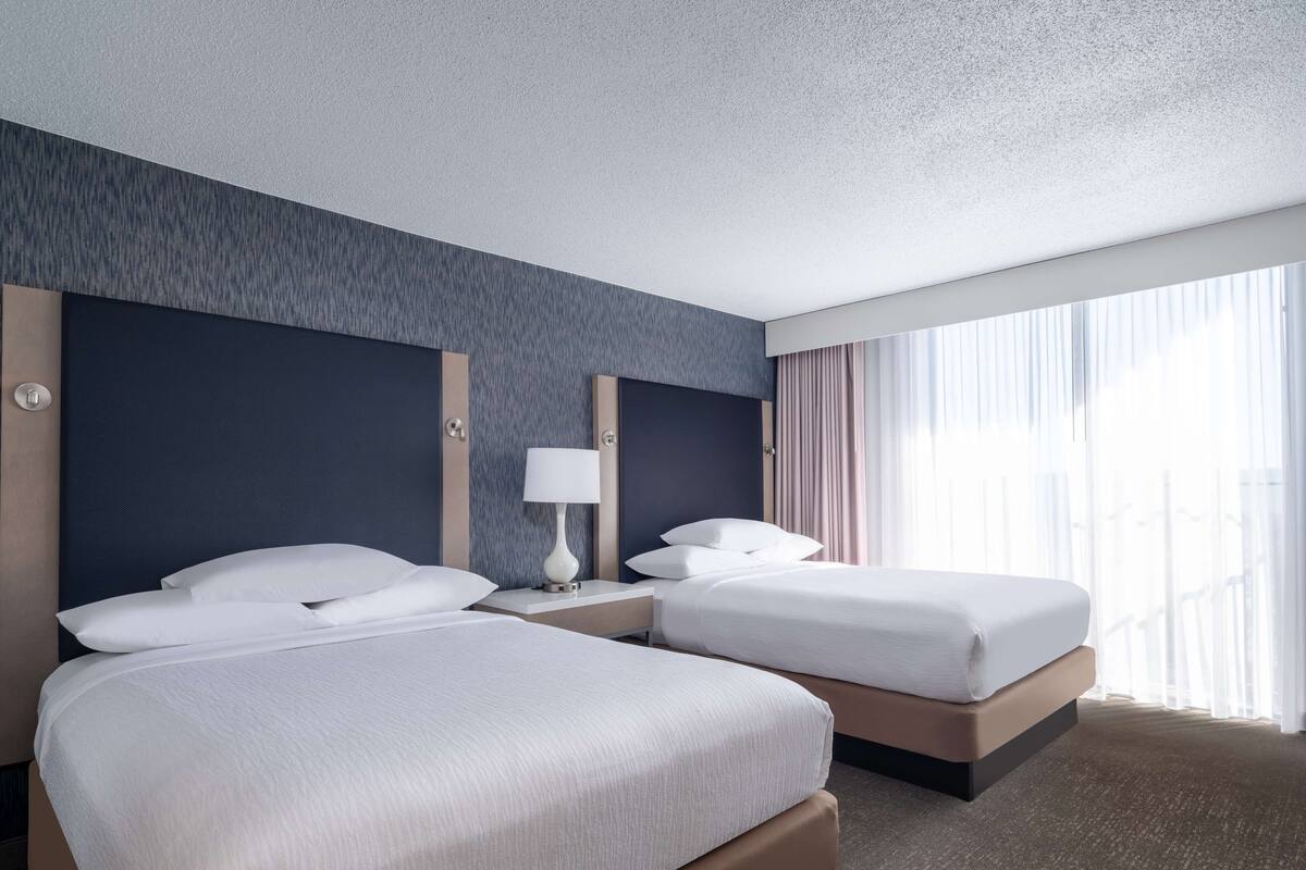 A hotel room with two beds, a nightstand with a lamp, a large window with curtains, and a textured wall.