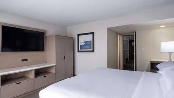 Habitación de hotel con cama, televisor en la pared, armario, escritorio con cajones, un cuadro enmarcado en la pared y una lámpara en la mesita de noche.