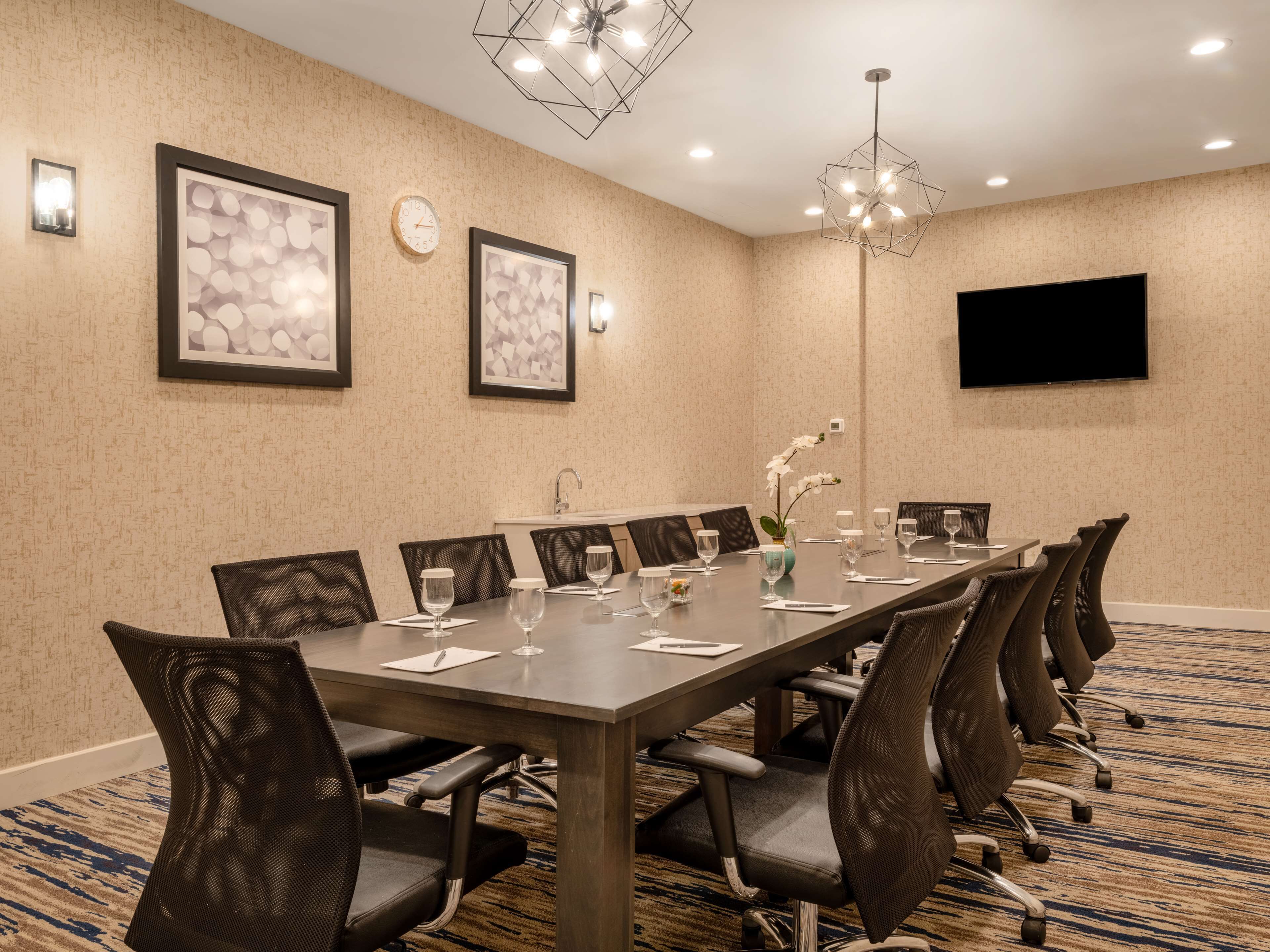 A conference room with a long table, chairs, a clock, framed artwork, a vase with flowers, and a TV on the wall.