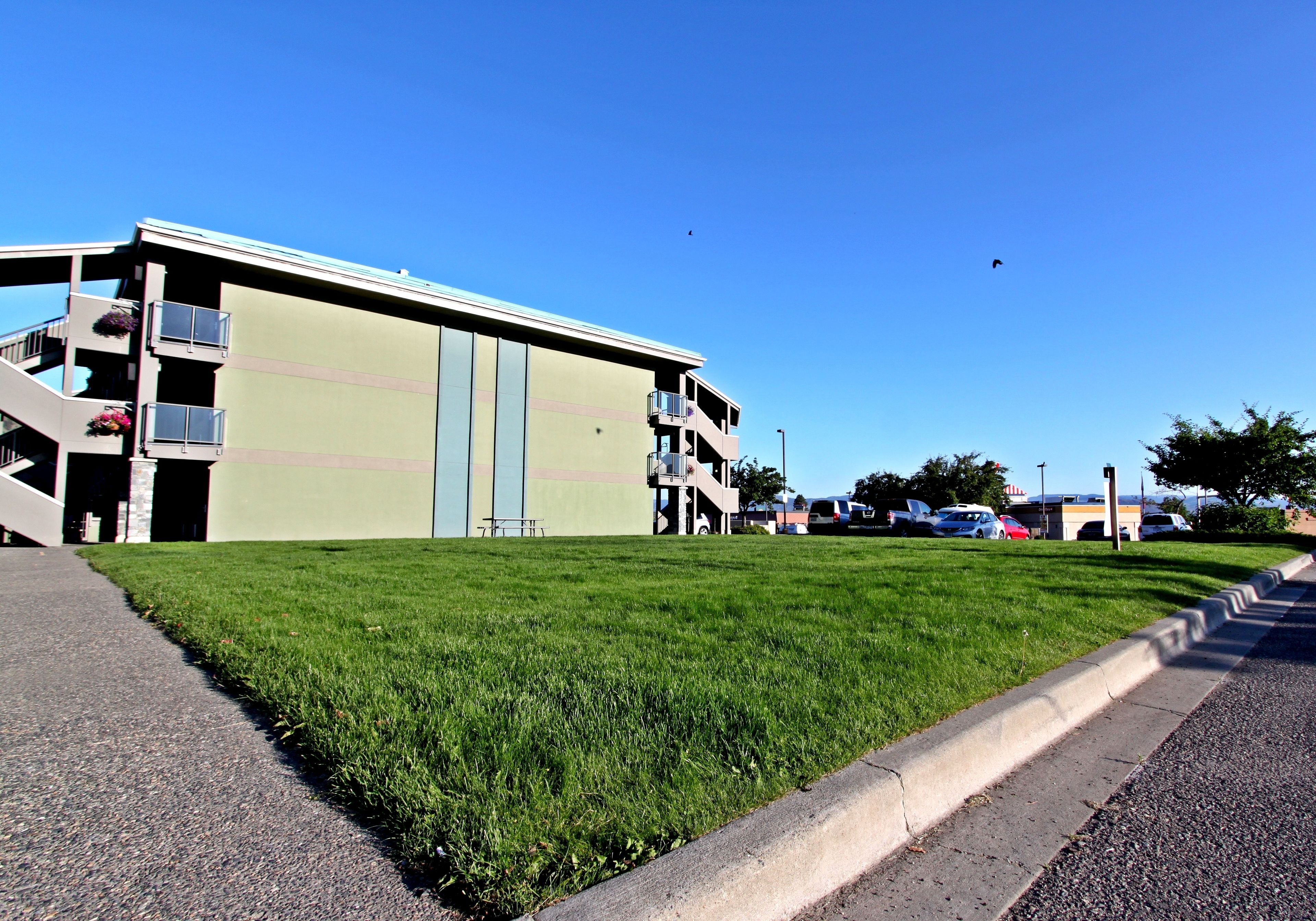 A multi-story building with a flat roof, a grassy lawn, and a parking lot with several cars.