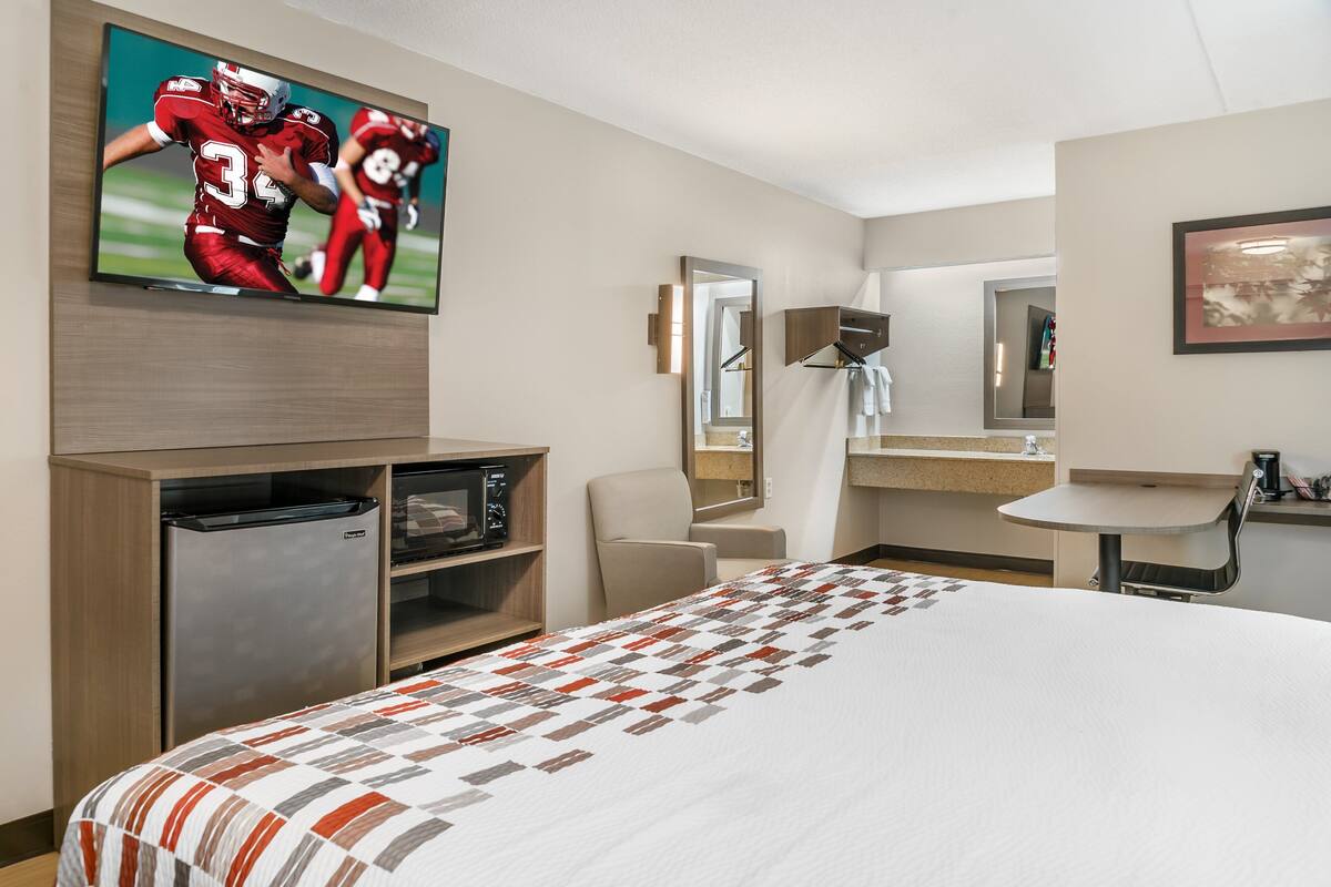 A hotel room with a large bed, a television displaying a football game, a minibar, a seating area, and a bathroom with a mirror and sink.