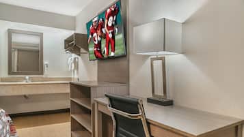 A hotel room with a wooden desk, a black chair, a television mounted on the wall, a mirror, and a lamp.