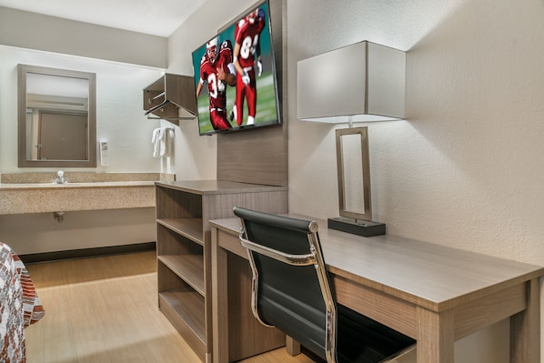 A hotel room with a wooden desk, a black chair, a television mounted on the wall, a mirror, and a lamp.