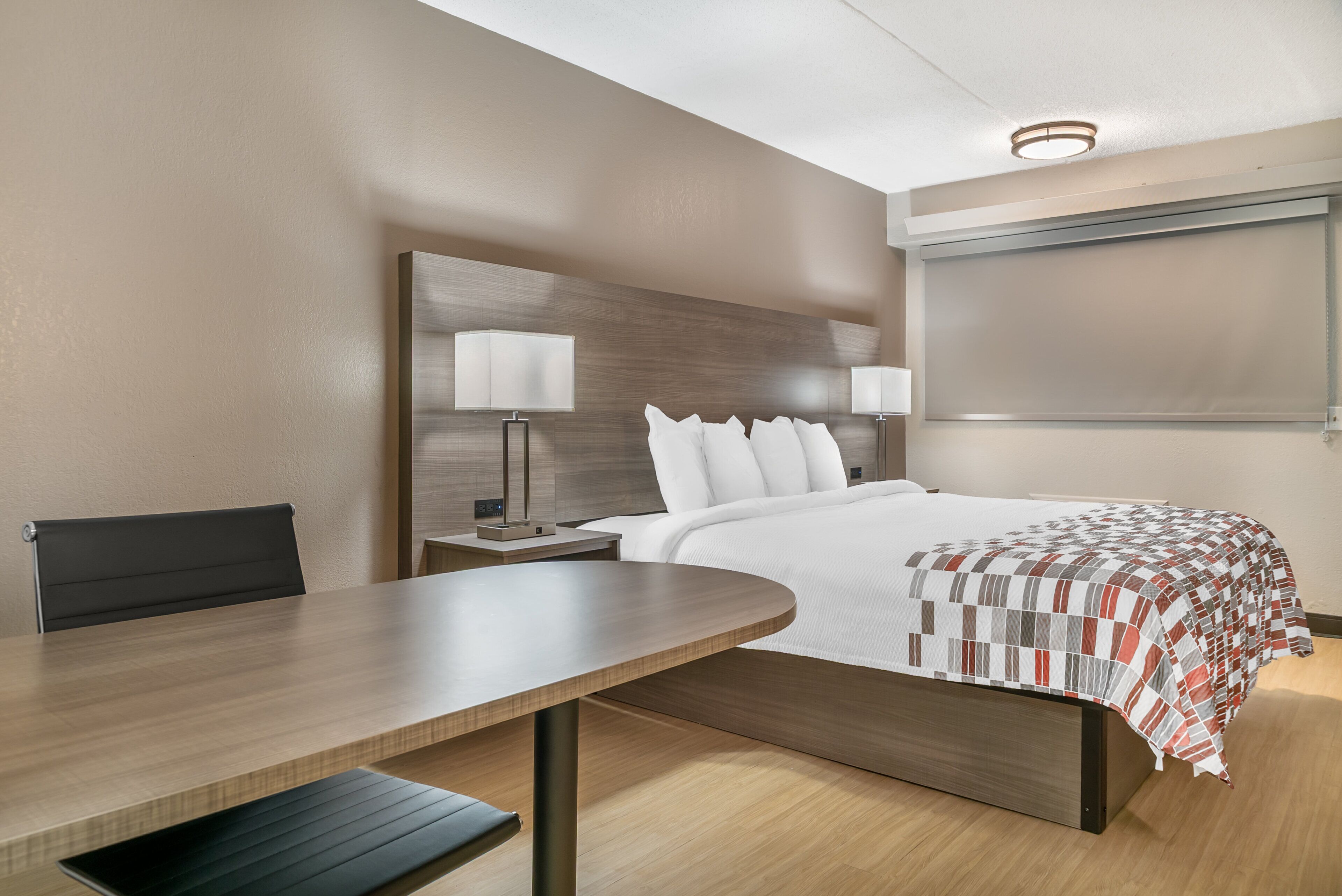 A modern hotel room with a wooden bed, bedside tables with lamps, a wooden desk, and a bench.