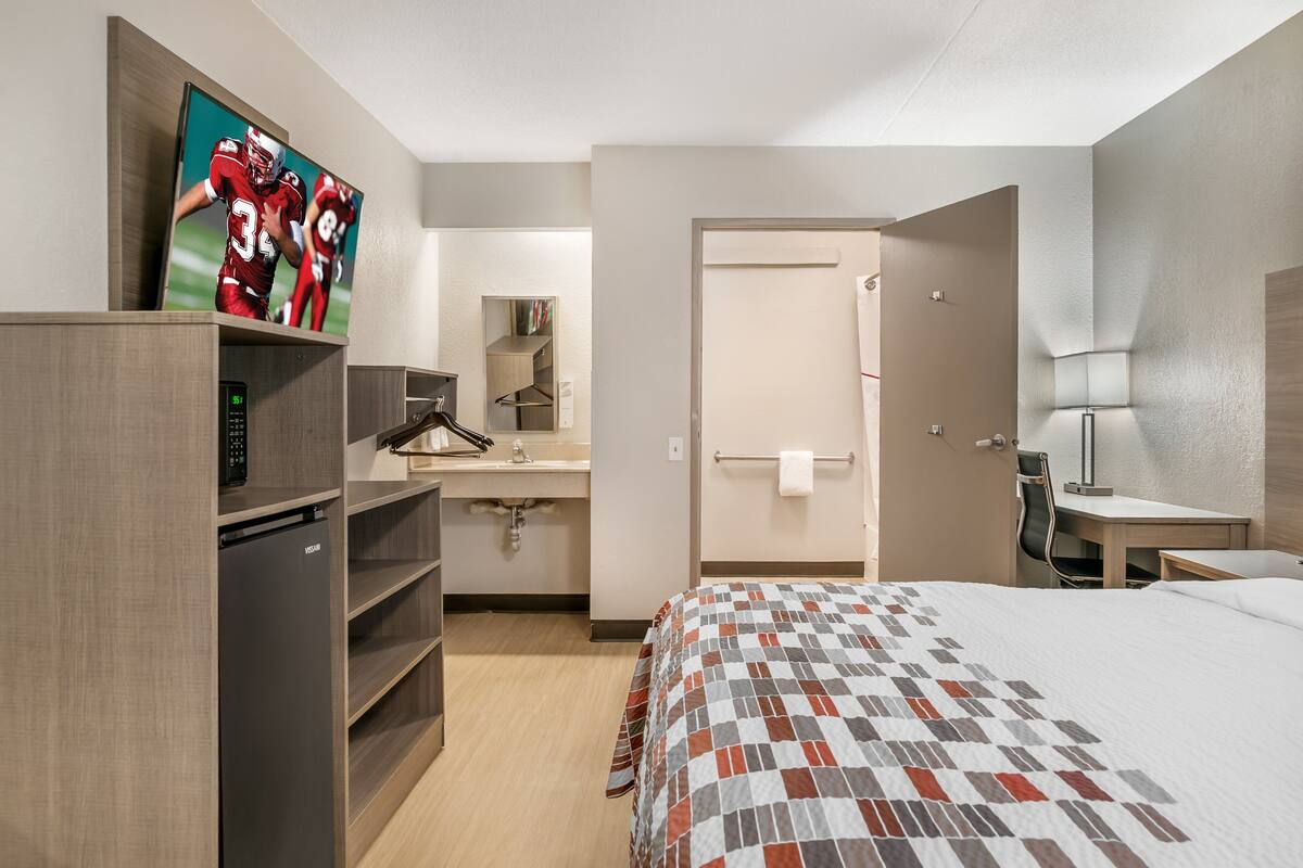 A hotel room with a bed, a desk, a chair, a TV, a minibar, a mirror, and a bathroom door.