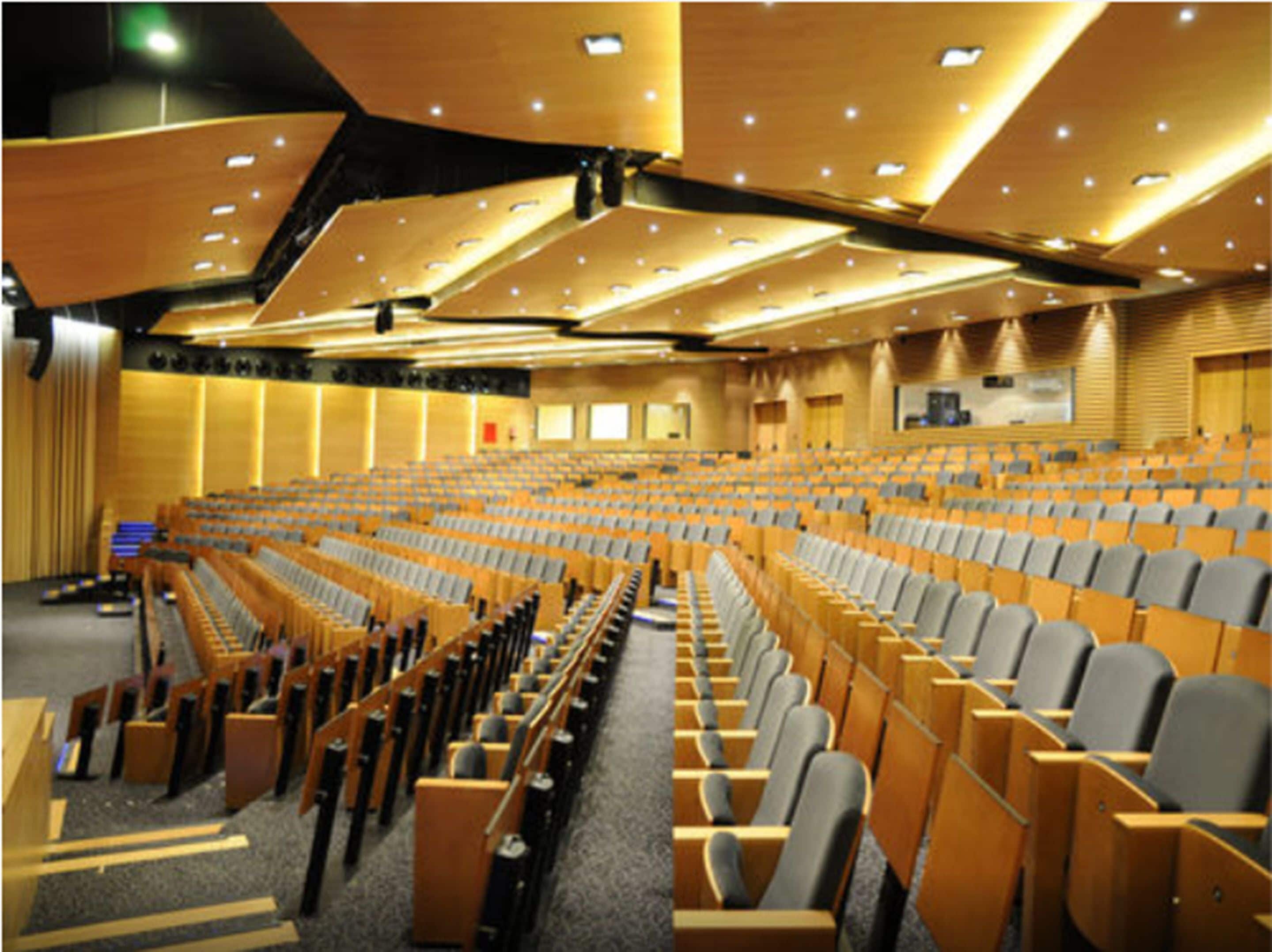Un amplio salón de conferencias con filas de sillas de madera y un escenario al frente.
