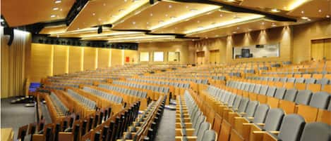 Un amplio salón de conferencias con filas de sillas de madera y un escenario al frente.