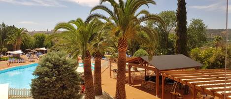 Zona de piscina amb gandules i una zona de seients coberta, envoltada de palmeres i amb vista a la ciutat al fons.
