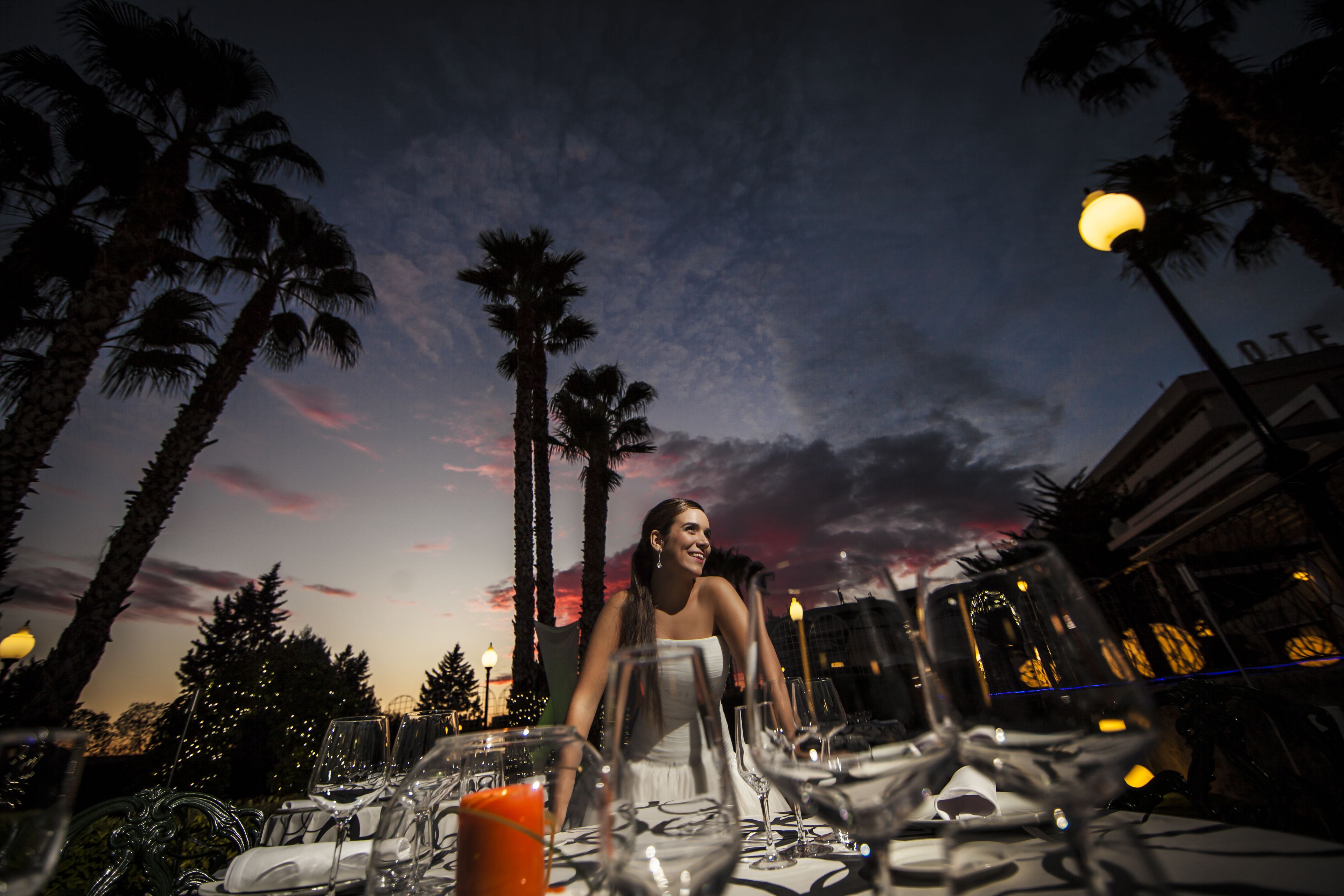 A table set for a meal with a sunset view, palm trees, and a building in the background.
