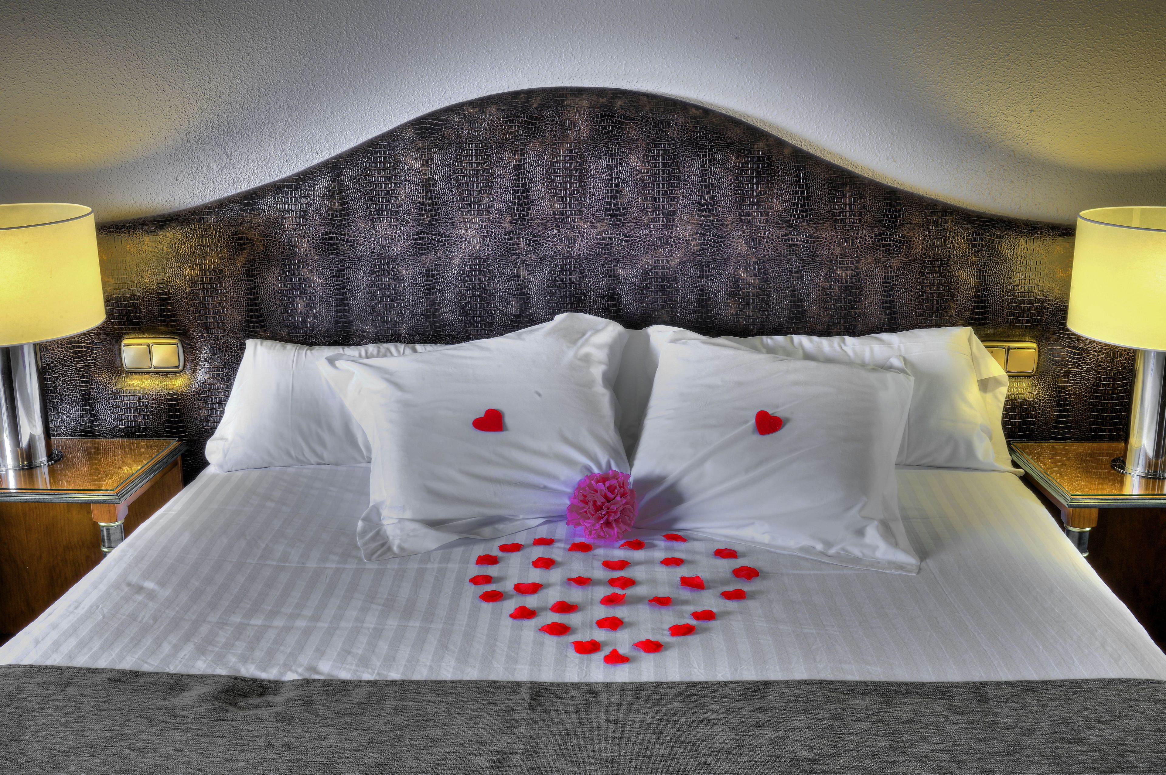 A neatly made bed with white linens, a decorative heart pattern on the bedspread, and two bedside lamps on wooden nightstands.