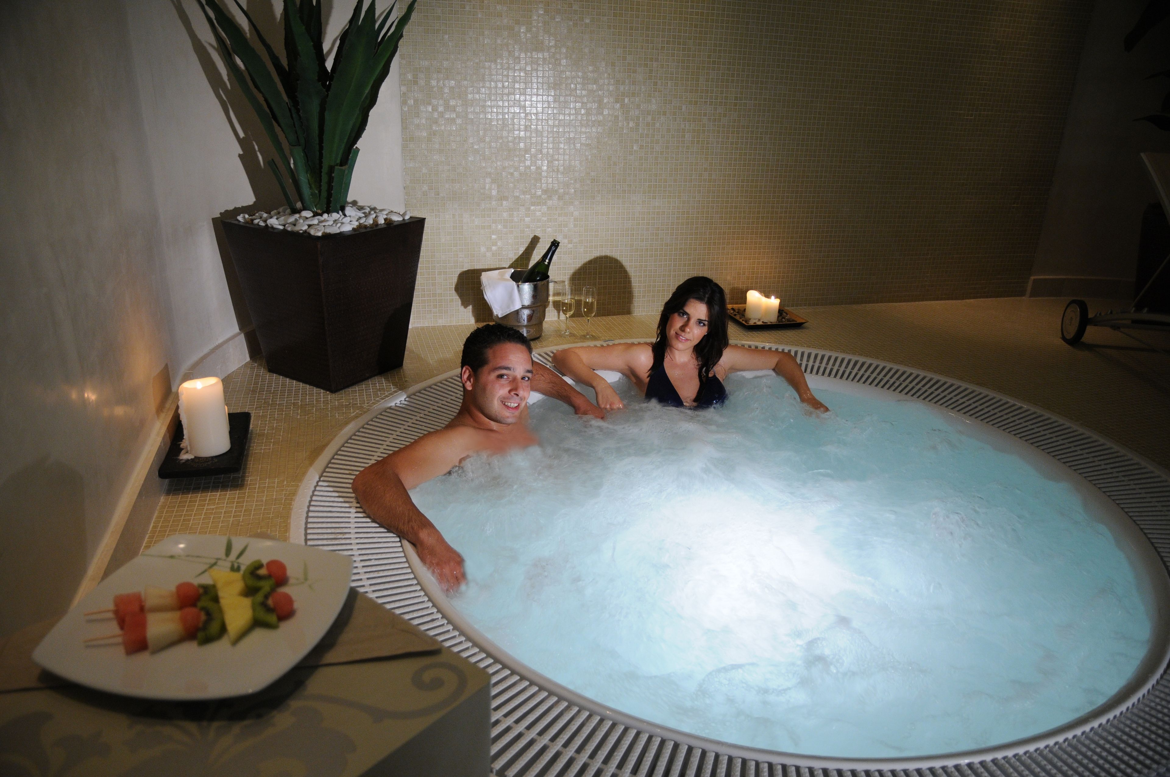 A couple in a hot tub with a candle, a potted plant, and a plate of fruit.