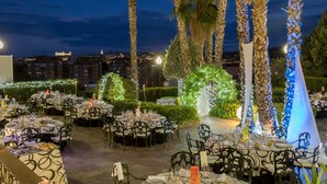An outdoor dining area with round tables and chairs, surrounded by palm trees and greenery, with city lights in the background. - Hotel Beatriz Toledo Auditorium & Spa (Toledo)
