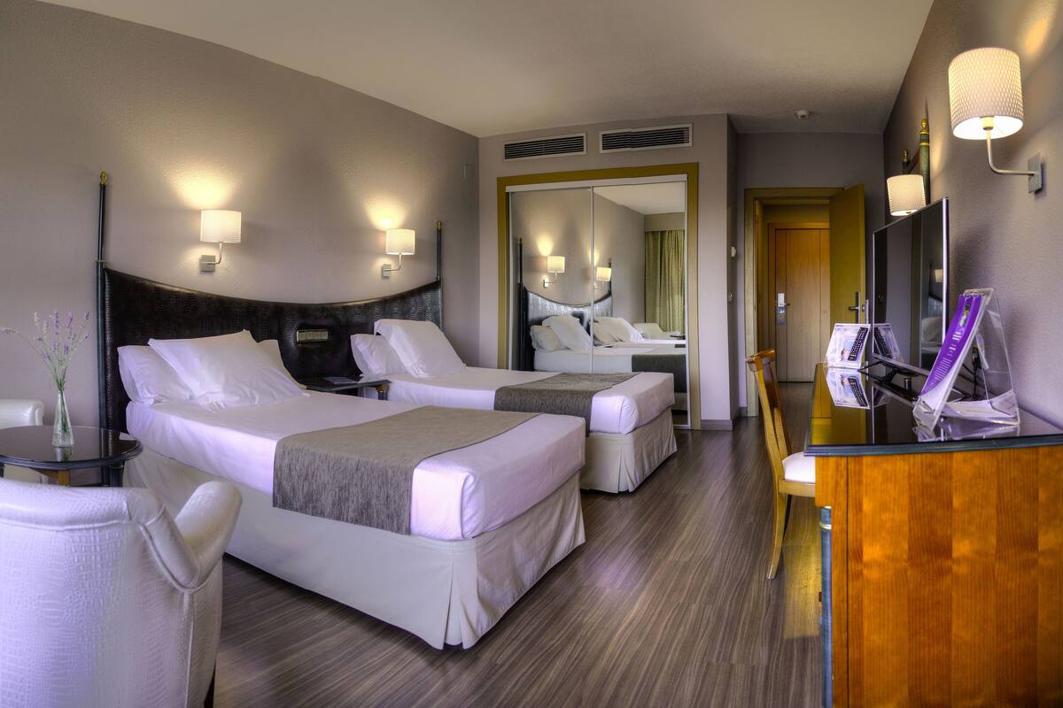 A hotel room with two beds, a desk, a chair, a lamp, and a mirror.