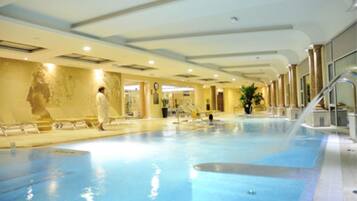 An indoor swimming pool with a person walking by, surrounded by a modern interior with a large wall mural and a staircase leading to an upper level.